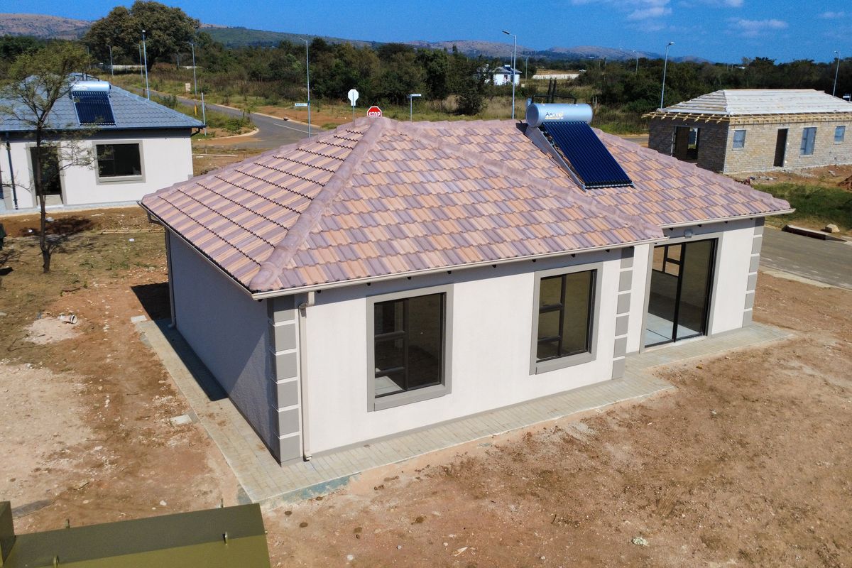 Aerial view of a modern single-story house featuring a tiled roof, solar water heater, and paved perimeter, set within an developing residential area.