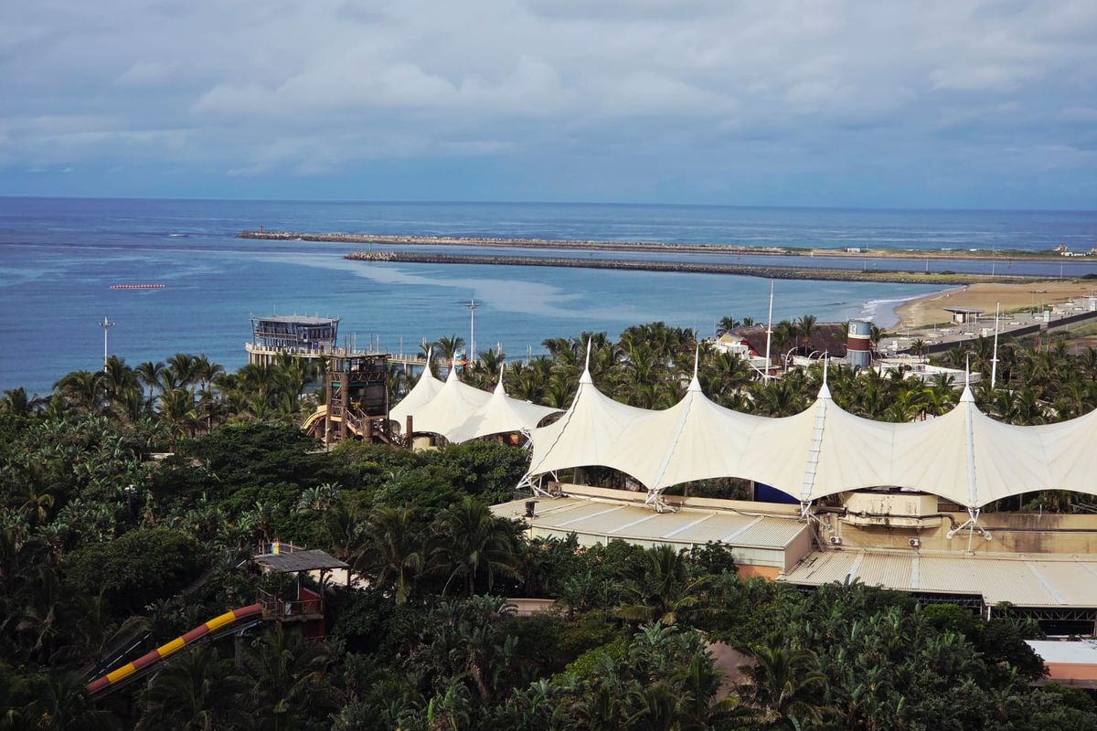 An aerial view of a coastal resort area featuring large tensile fabric roof structures, lush tropical vegetation, and a water slide near the ocean.