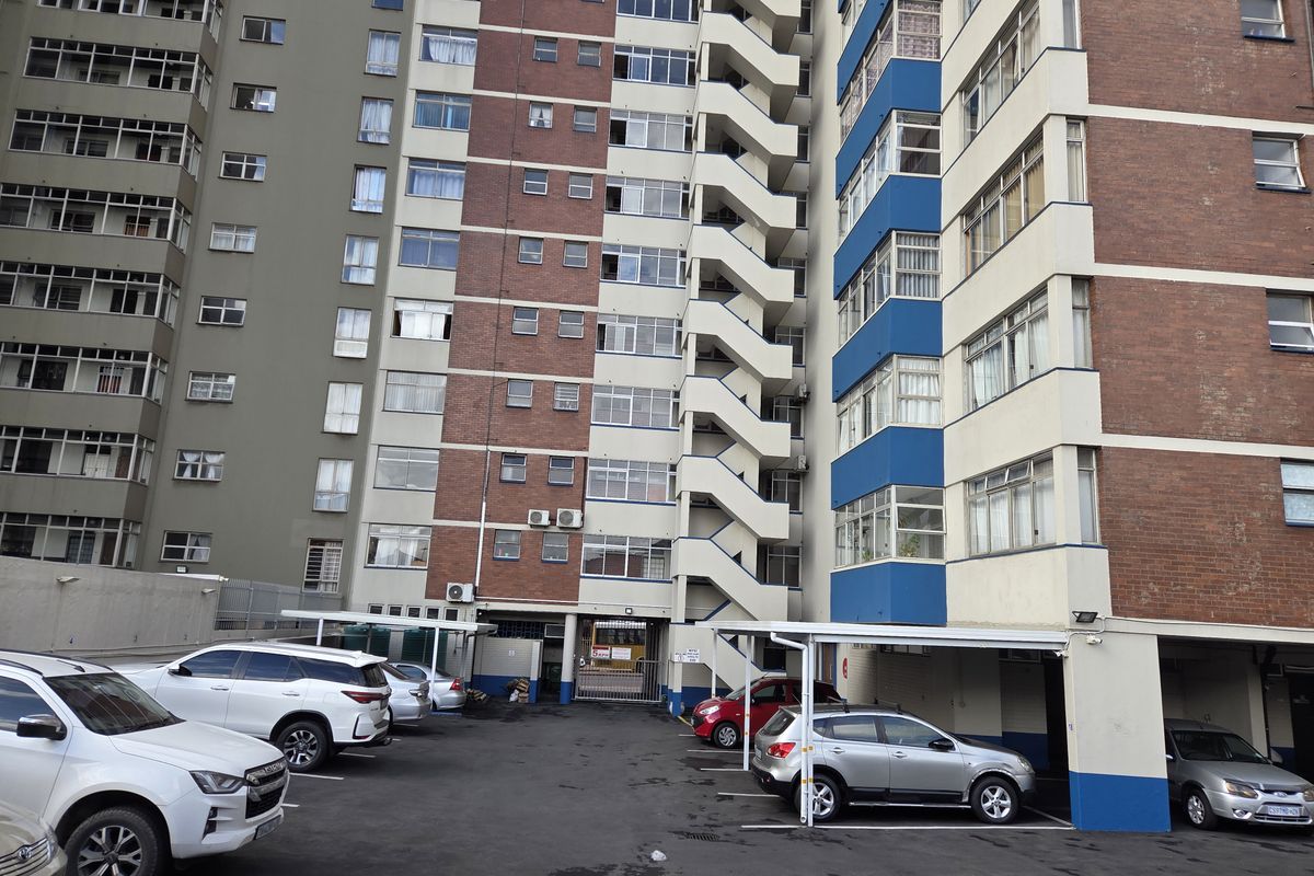 Rear view of a multi-story apartment building featuring an asphalt parking area with covered carports and an external staircase.