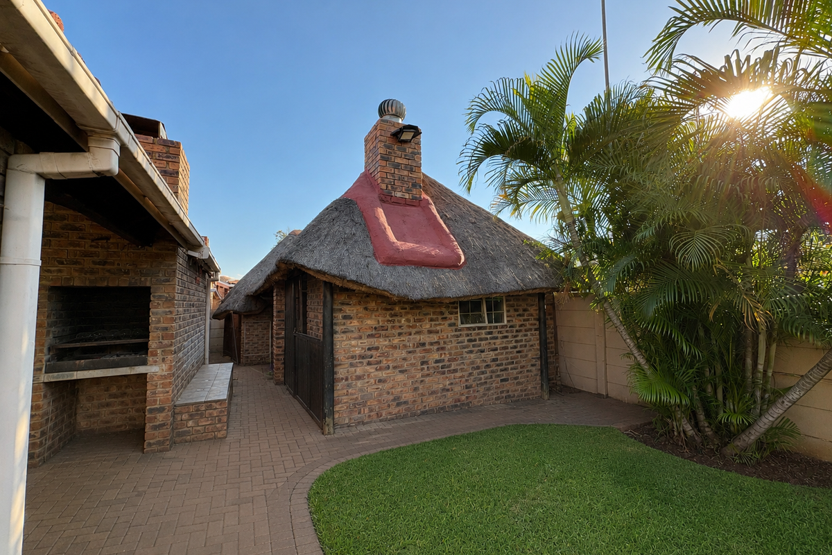 Rear garden area featuring a thatched lapa with a built-in braai, paved patio, and established palm trees.