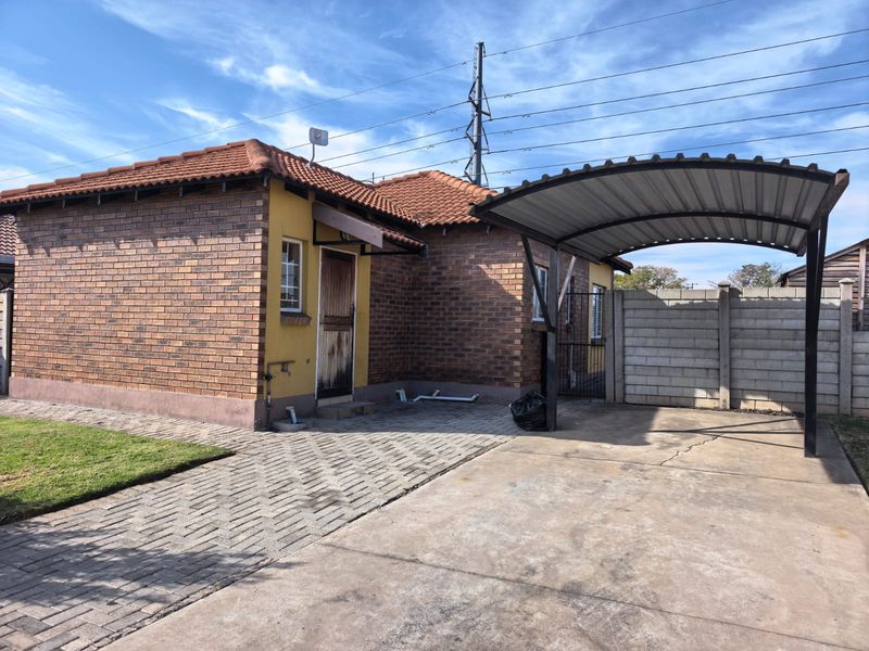 Front exterior view of a brick house featuring a tiled roof, a covered carport, and a paved driveway with a precast boundary wall.