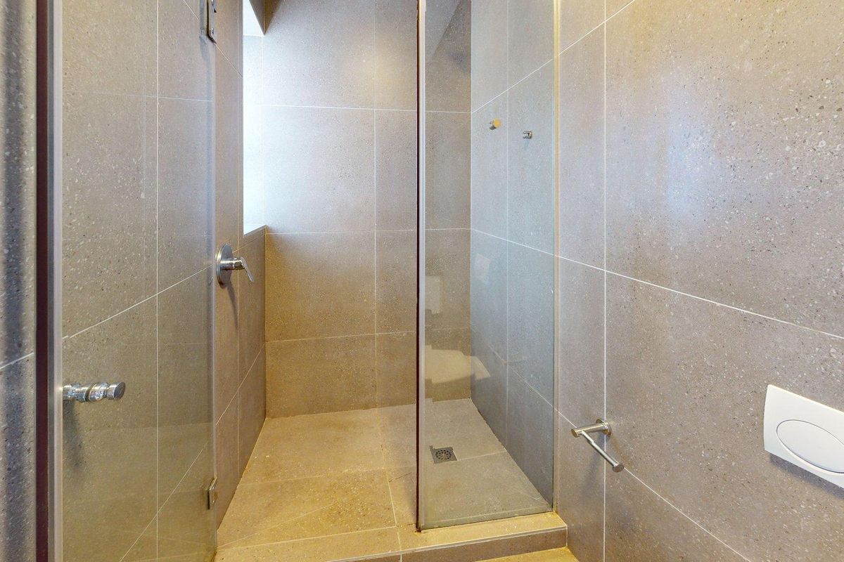 A modern bathroom featuring a walk-in shower with a glass screen, neutral stone-look wall and floor tiling, and a wall-mounted flush plate.