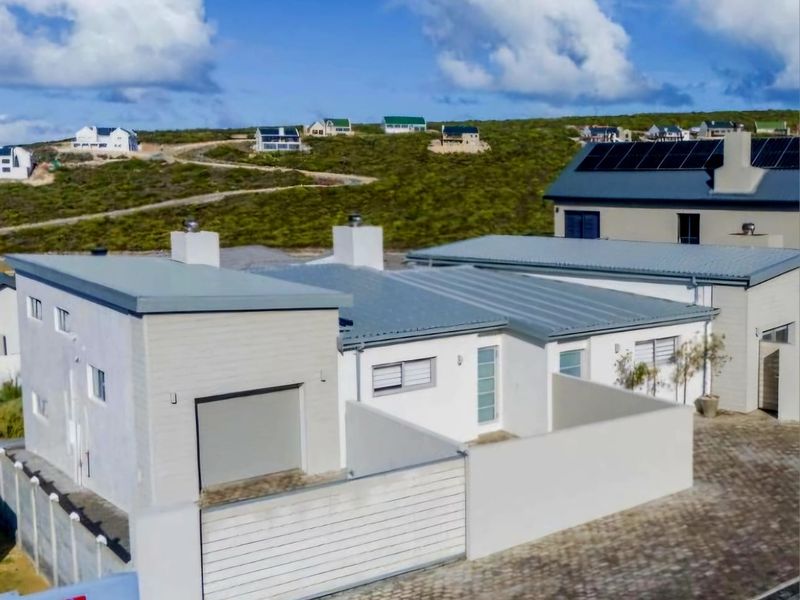 Aerial view of a modern residential property featuring a metal roof, solar panels, paved driveway, and secure boundary walling in a coastal estate setting.