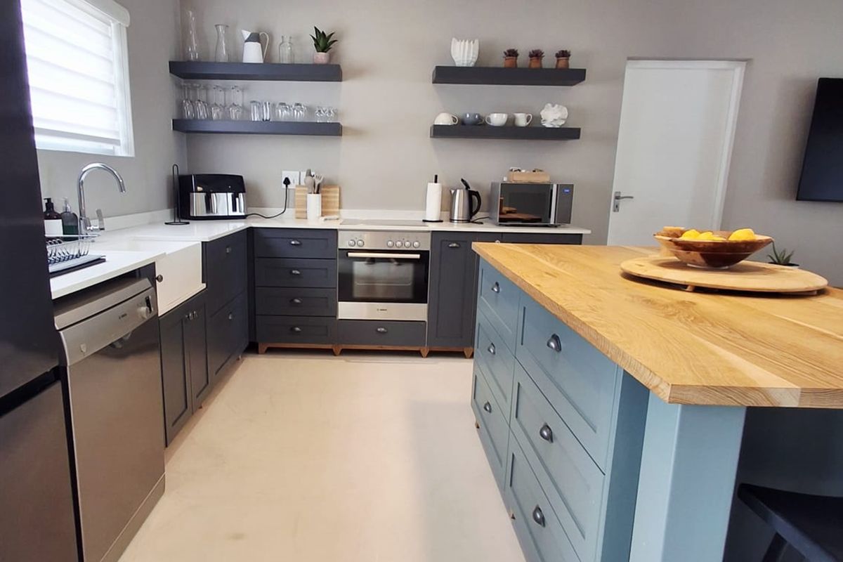 Modern kitchen featuring dark cabinetry, a large timber-topped island, apron sink, integrated oven, and floating shelving with contemporary pendant lighting.