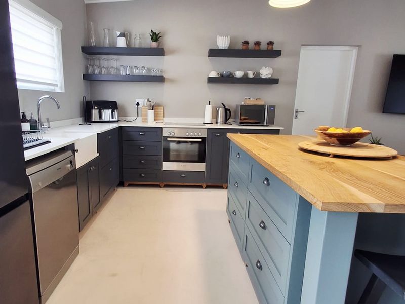 Modern kitchen featuring dark cabinetry, a large timber-topped island, apron sink, integrated oven, and floating shelving with contemporary pendant lighting.