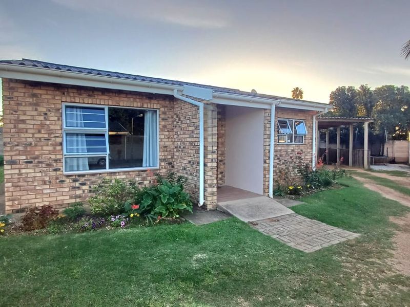 A well-maintained face brick townhouse exterior featuring a covered entrance, paved walkway, and established garden beds under a tiled roof.