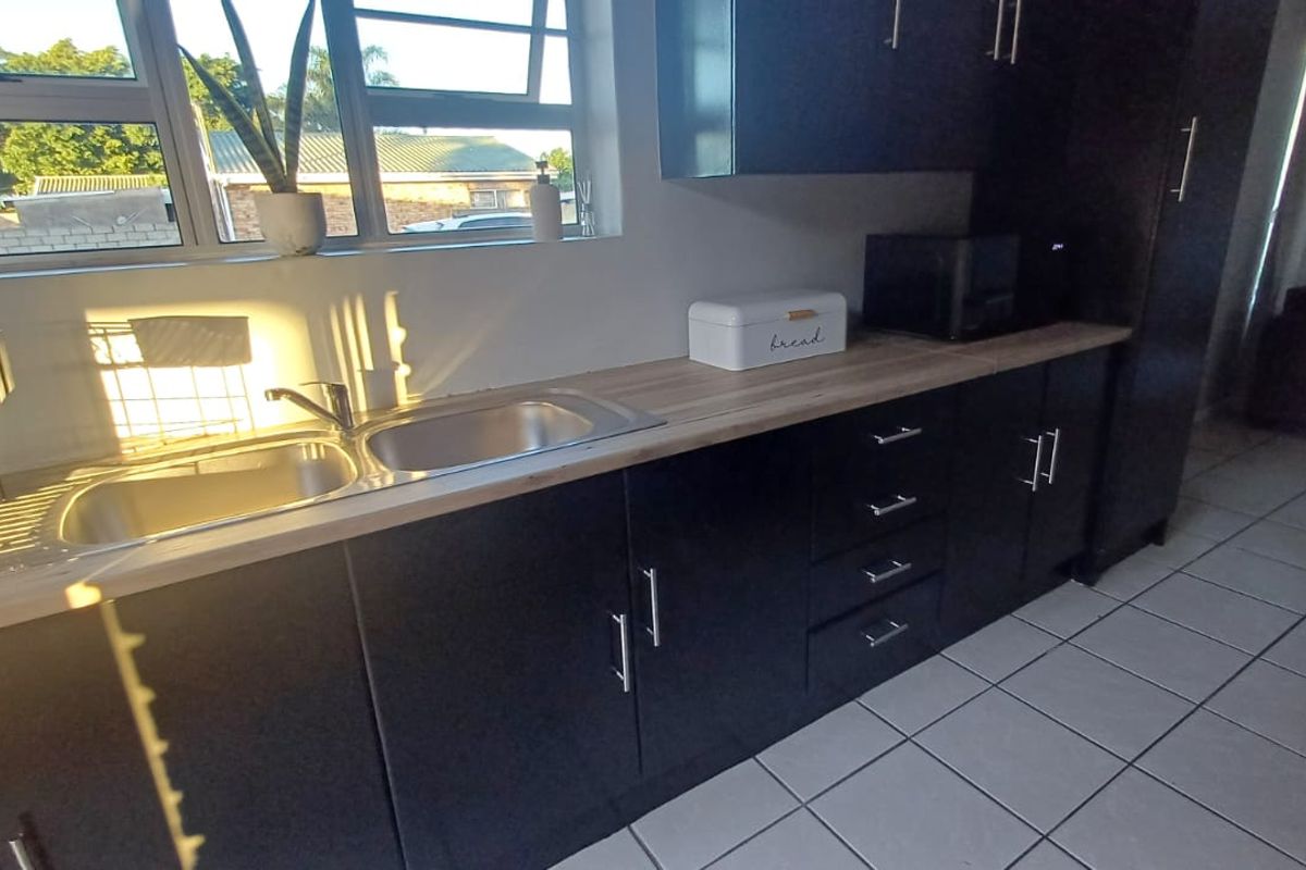 A modern kitchen featuring dark cabinetry, a light-toned laminate countertop, a double bowl stainless steel sink, and tiled flooring with natural light.