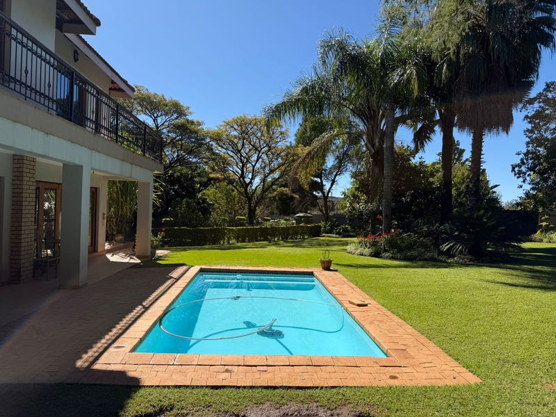 A well-maintained swimming pool set within a lush, landscaped garden, featuring a covered patio and an upper-level balcony with wrought iron railings.