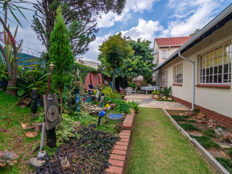 A rear garden view featuring a paved patio area, established landscaping, brick garden borders, and the rear elevation of the house with security window bars.