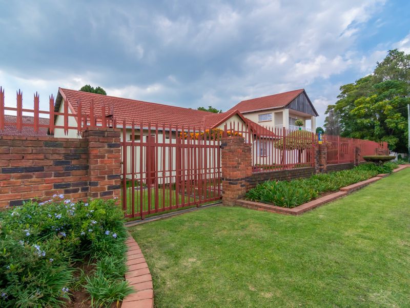 Front view of the property featuring a well-maintained garden, brick boundary wall, and secure metal fencing with a tiled roof house in the background.