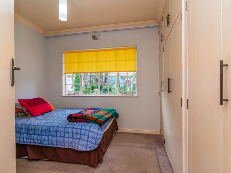 A functional bedroom featuring built-in cupboards, neutral wall finishes, and a window with burglar bars overlooking the garden.