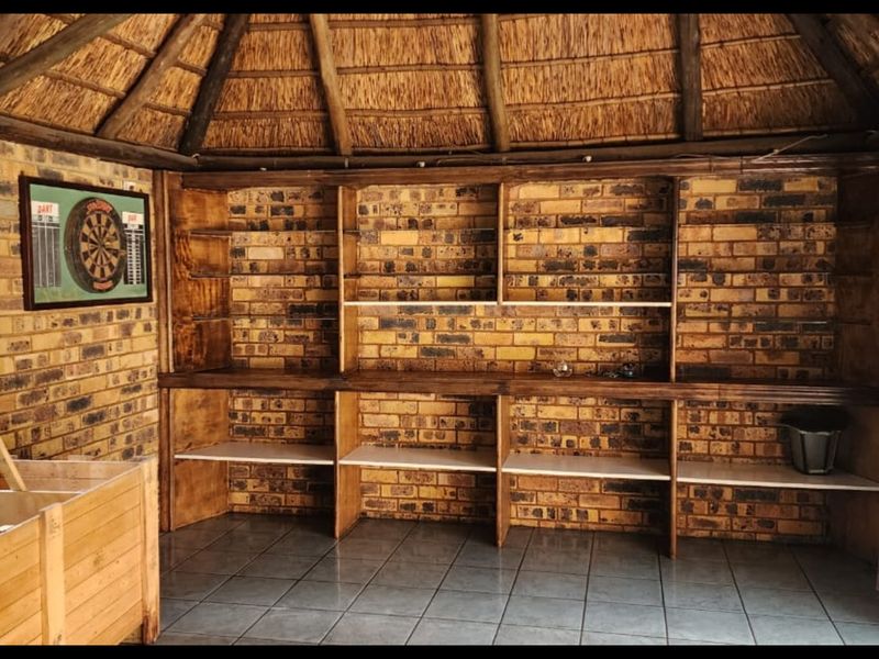 A rustic covered patio area featuring a traditional thatched roof, exposed timber rafters, facebrick walls, and built-in shelving with tiled flooring.