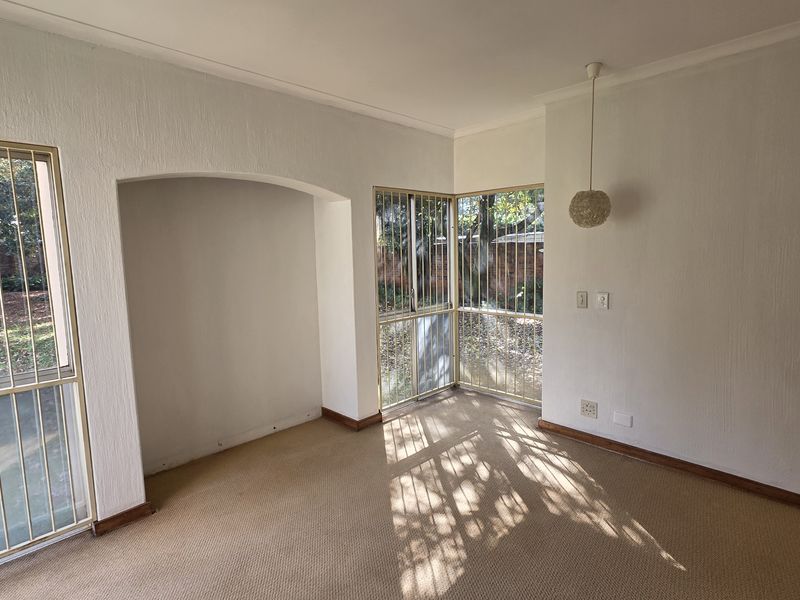 A bright, empty living area featuring an arched alcove, carpeted flooring, and large windows fitted with security bars overlooking the garden.
