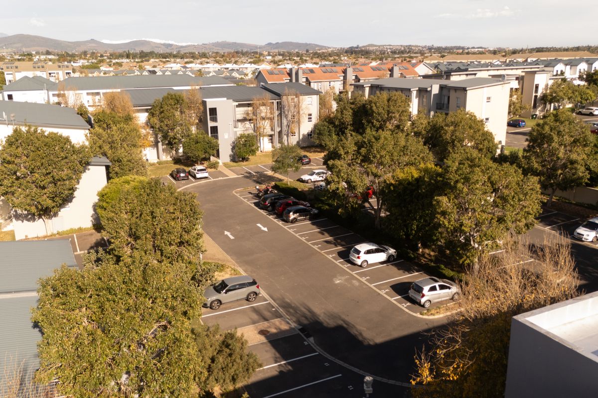 An elevated aerial view of the apartment complex grounds, featuring paved parking bays, mature trees, and surrounding residential building blocks.