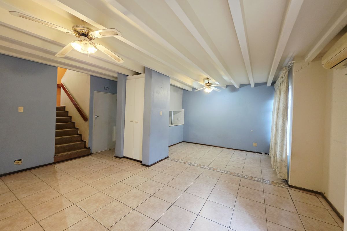Spacious open-plan living area featuring tiled floors, exposed beam ceilings, dual ceiling fans, and a staircase leading to the upper level.