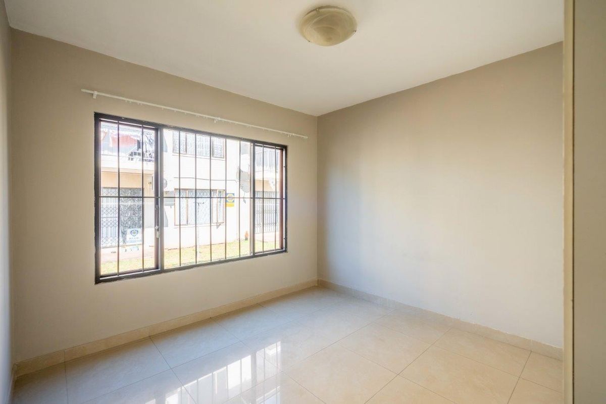 A bright, empty bedroom featuring neutral wall paint, tiled flooring, and a large window fitted with security bars.