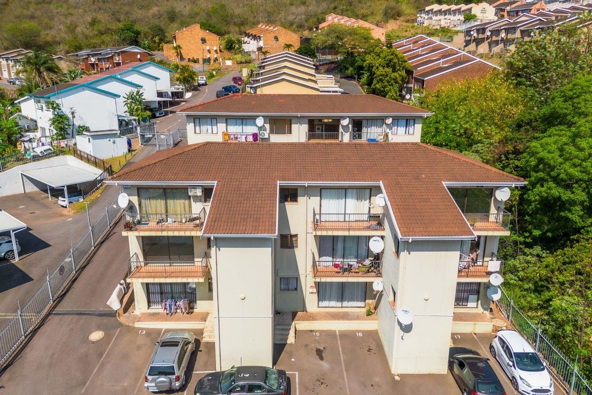 Aerial view of a multi-story townhouse complex featuring tiled roofing, private balconies, and an adjacent paved parking area within a fenced estate.