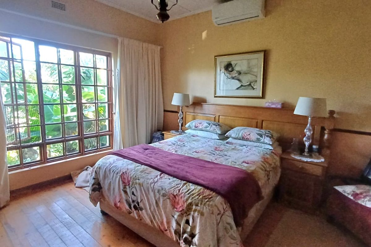 A cozy bedroom featuring warm timber flooring, a built-in headboard, and a split-system air conditioner, with large windows overlooking the garden.
