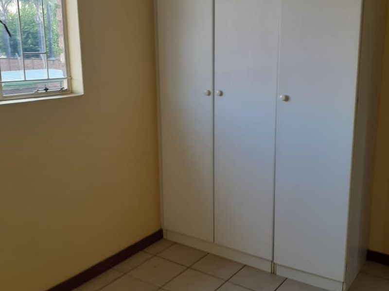 A simple, empty bedroom featuring tiled flooring, built-in cupboards, and a window with security bars.