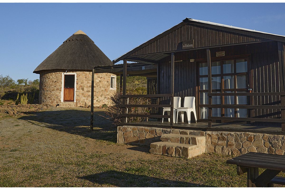 Exterior view of a rustic farm property featuring a traditional thatched-roof stone rondavel and a timber-clad cabin with a raised wooden deck.