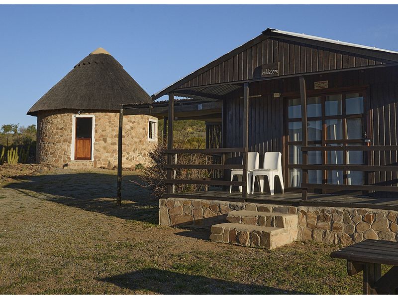 Exterior view of a rustic farm property featuring a traditional thatched-roof stone rondavel and a timber-clad cabin with a raised wooden deck.