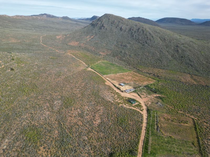 Aerial view of a rural farm property featuring expansive landscape, a dirt access road, fenced paddocks, and existing outbuildings.