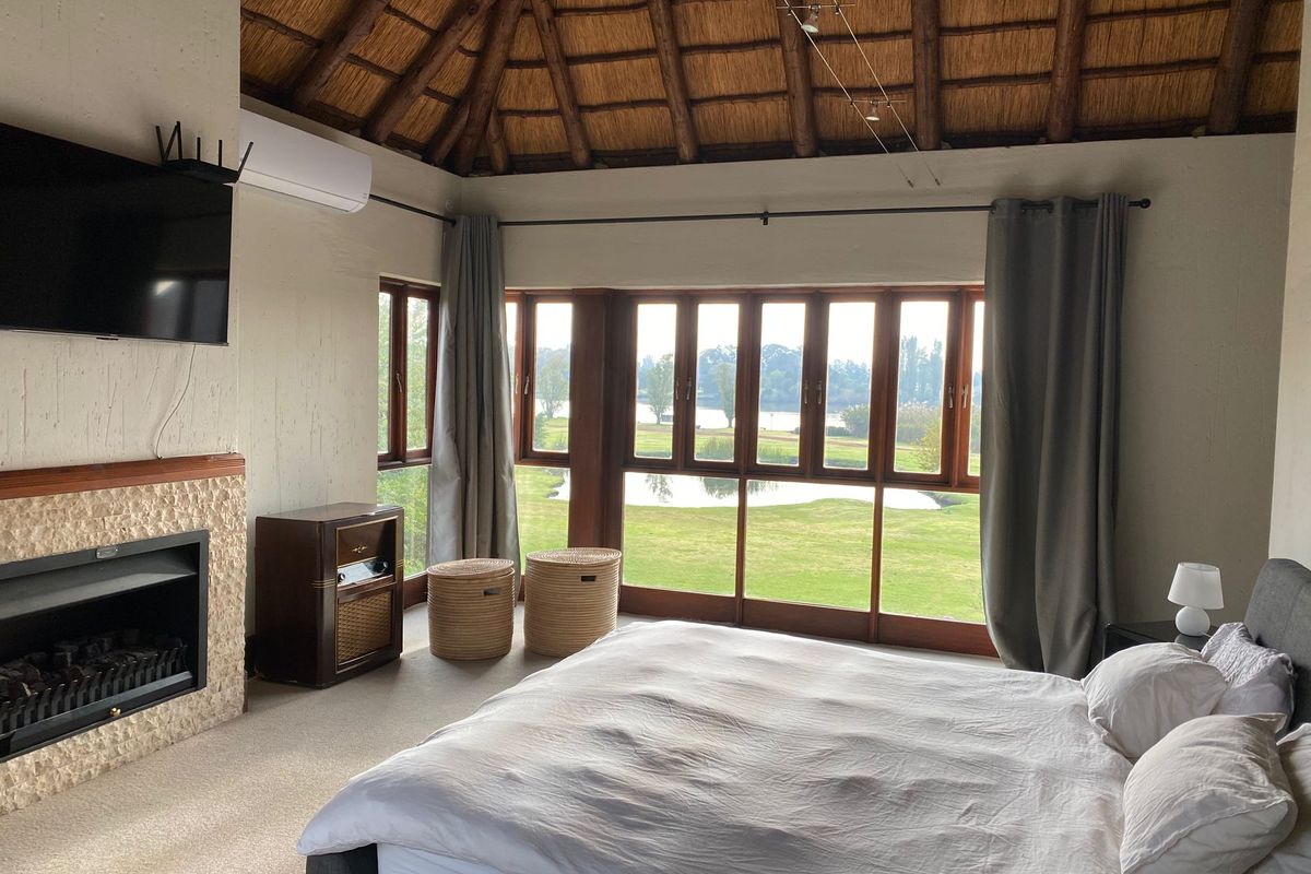 Spacious bedroom featuring a high thatched ceiling with exposed rafters, a built-in stone fireplace, and large windows offering scenic golf course and water views.
