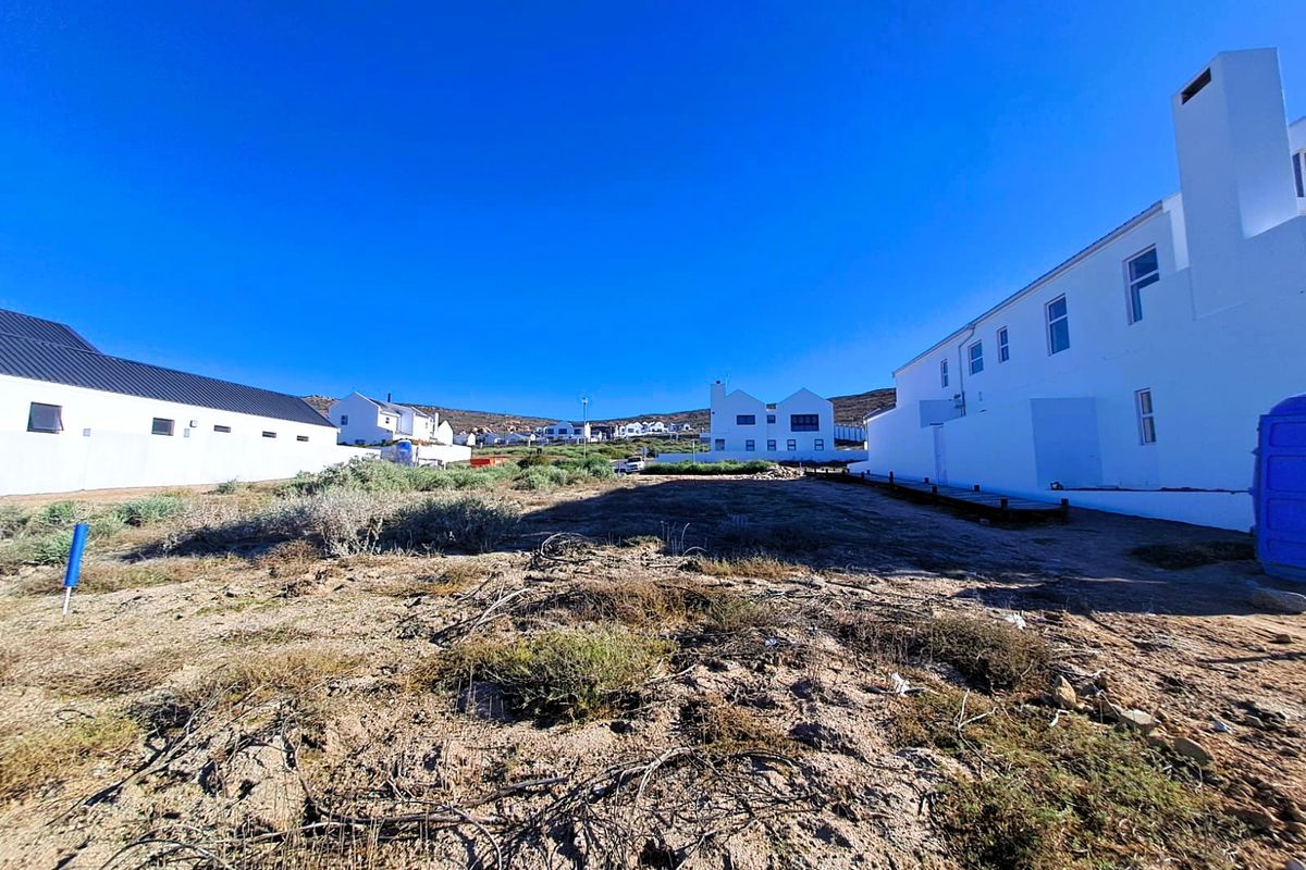 A view of a vacant residential plot situated between existing white-walled residential properties under a clear blue sky.
