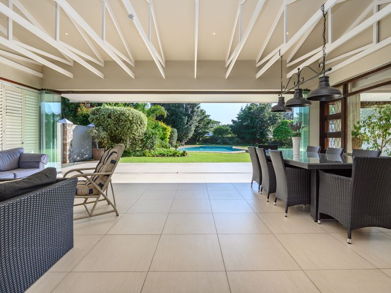 Spacious covered patio featuring exposed white rafters, large format tiling, and seamless stacking glass doors opening to a landscaped garden and swimming pool area.