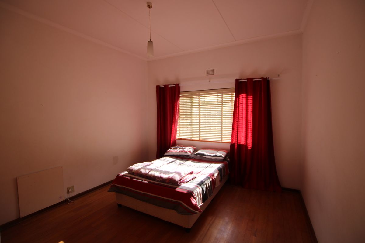 A bedroom featuring warm wooden flooring, a window with venetian blinds, and a ceiling light fixture.