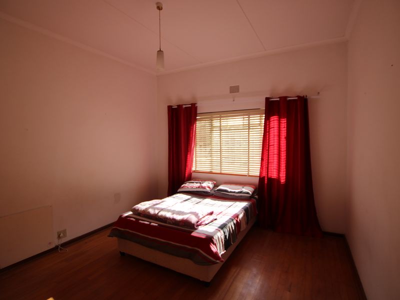 A bedroom featuring warm wooden flooring, a window with venetian blinds, and a ceiling light fixture.