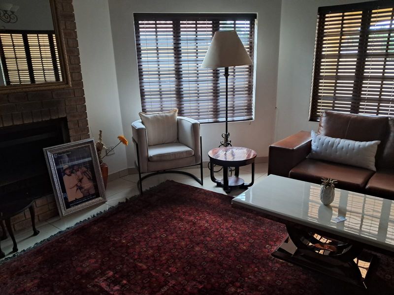 A cozy living room featuring a brick fireplace, tiled flooring, and large windows fitted with venetian blinds.