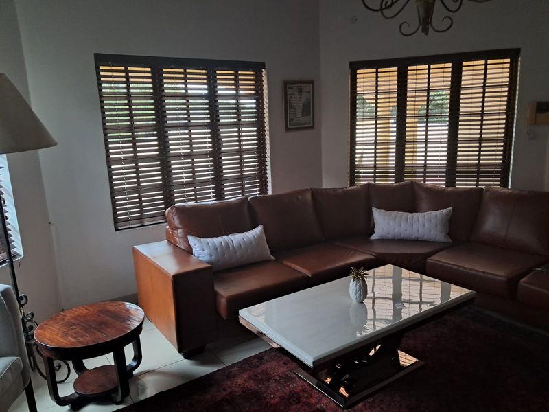 A comfortable living room featuring tiled flooring, large windows with wooden venetian blinds, and a decorative wrought iron chandelier.
