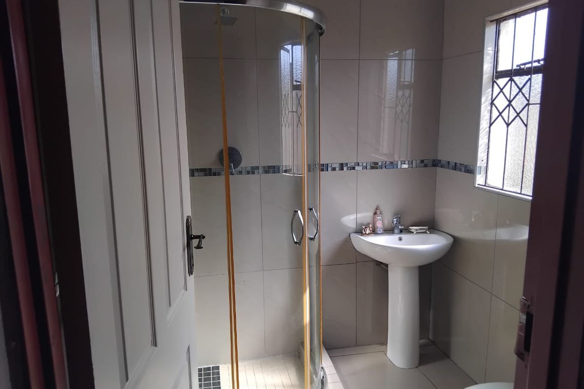 A functional bathroom featuring a curved glass shower enclosure, pedestal basin, and neutral wall tiling with a decorative mosaic accent strip.