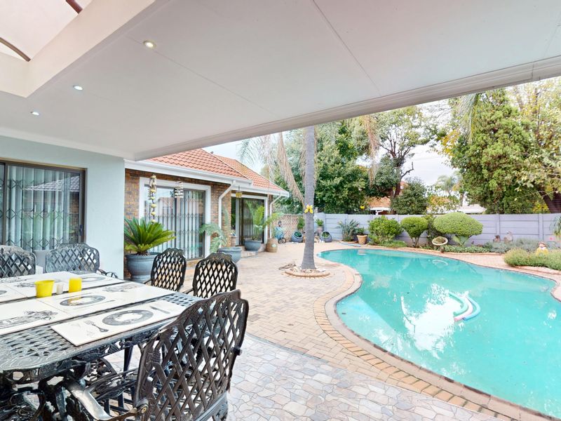 A spacious covered patio overlooking a private swimming pool and landscaped garden, featuring paved flooring and direct access to the home via sliding doors.