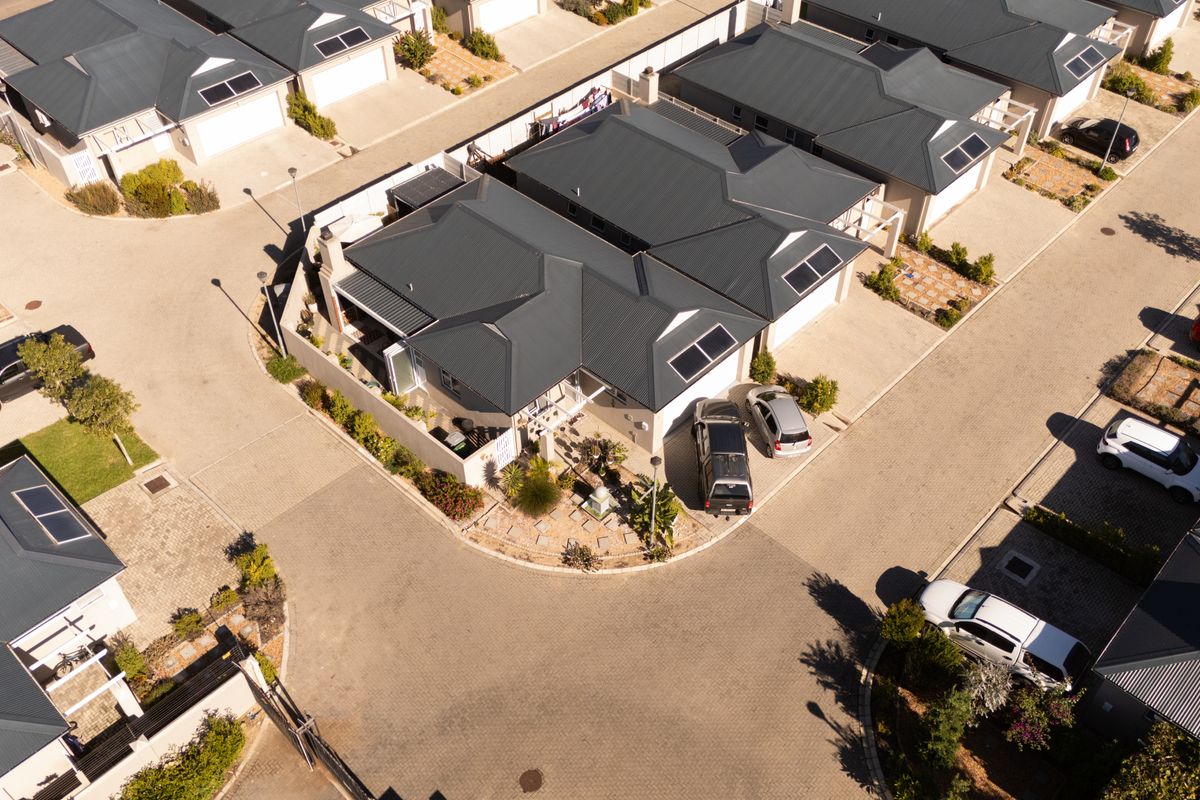 Aerial view of a residential property featuring a metal roof with solar panels, a paved driveway, and surrounding neighborhood infrastructure.