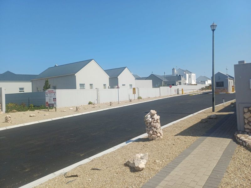 A view of a paved private road within a residential estate, featuring modern boundary walls, street lighting, and a security gatehouse.
