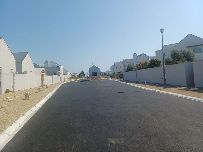 A view of a paved residential street within a secure estate, featuring a central security gatehouse and perimeter boundary walls.