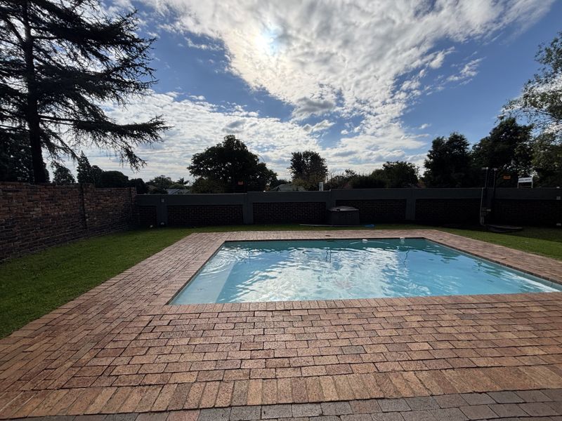 A well-maintained outdoor swimming pool area featuring brick paving, a surrounding lawn, and a secure perimeter brick wall.