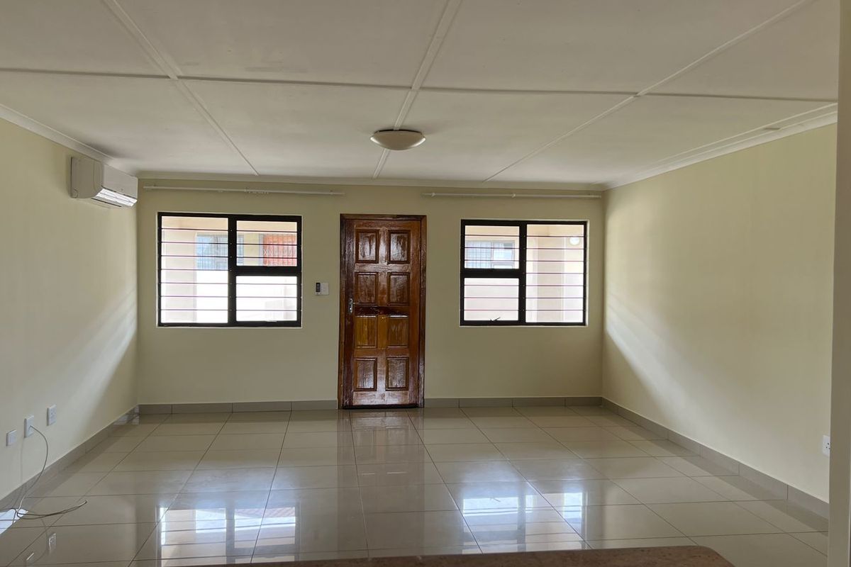 A spacious, empty living area featuring neutral tiled flooring, a split-system air conditioner, and secure windows with fitted security bars.