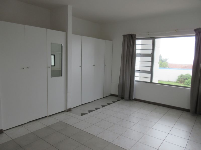 A spacious, empty bedroom featuring extensive built-in cupboards, neutral tiled flooring, and a large window providing natural light.