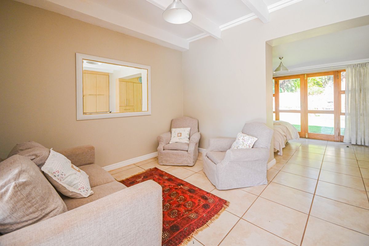 A bright living area featuring tiled floors, exposed ceiling beams, and an interior window looking into the kitchen, with access to the garden.