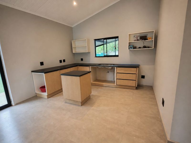 A bright, open-plan kitchen featuring light wood cabinetry, black countertops, a stainless steel sink, and vaulted ceilings, currently awaiting appliance installation.