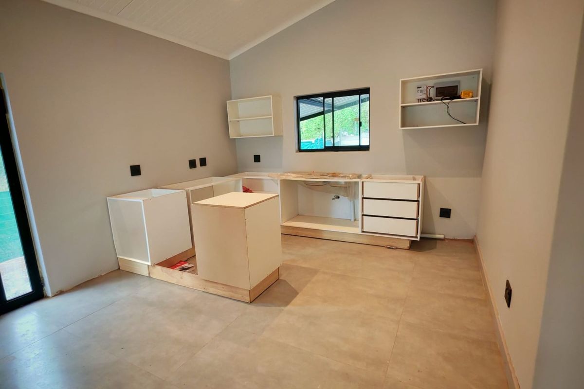 An unfinished kitchen space featuring installed base cabinetry, wall-mounted shelving, and tiled flooring, currently awaiting countertop and sink installation.