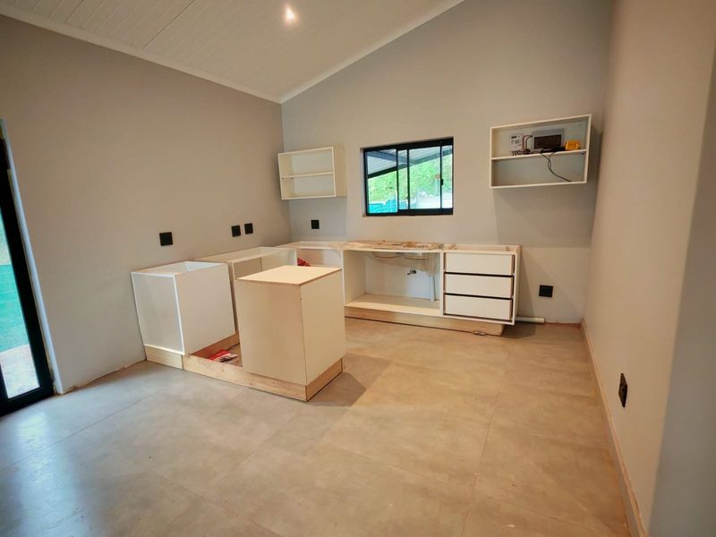 An unfinished kitchen space featuring installed base cabinetry, wall-mounted shelving, and tiled flooring, currently awaiting countertop and sink installation.