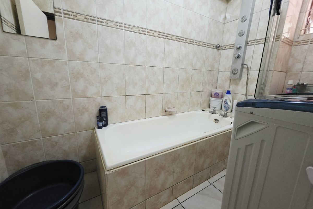 A functional bathroom featuring a built-in bathtub with a shower panel, tiled walls, and tiled flooring, requiring some cosmetic updates to refresh the space.