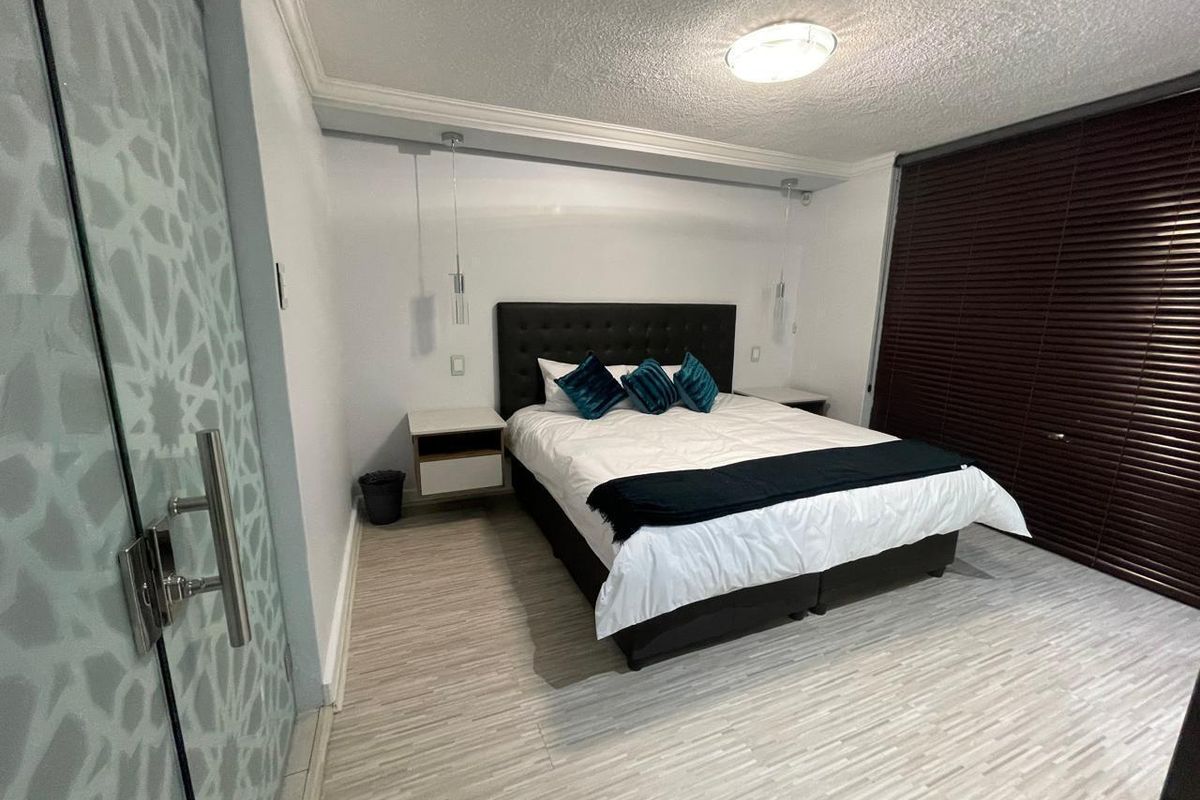 A well-maintained bedroom featuring modern laminate flooring, crown molding, built-in bedside tables with pendant lighting, and dark venetian blinds for privacy.