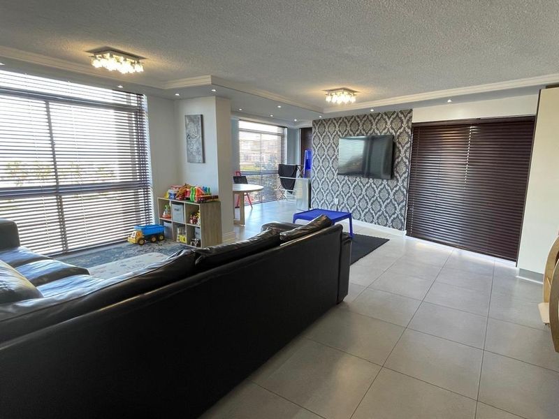 Spacious open-plan living area featuring large windows with venetian blinds, tiled flooring, recessed lighting, and a decorative feature wall.