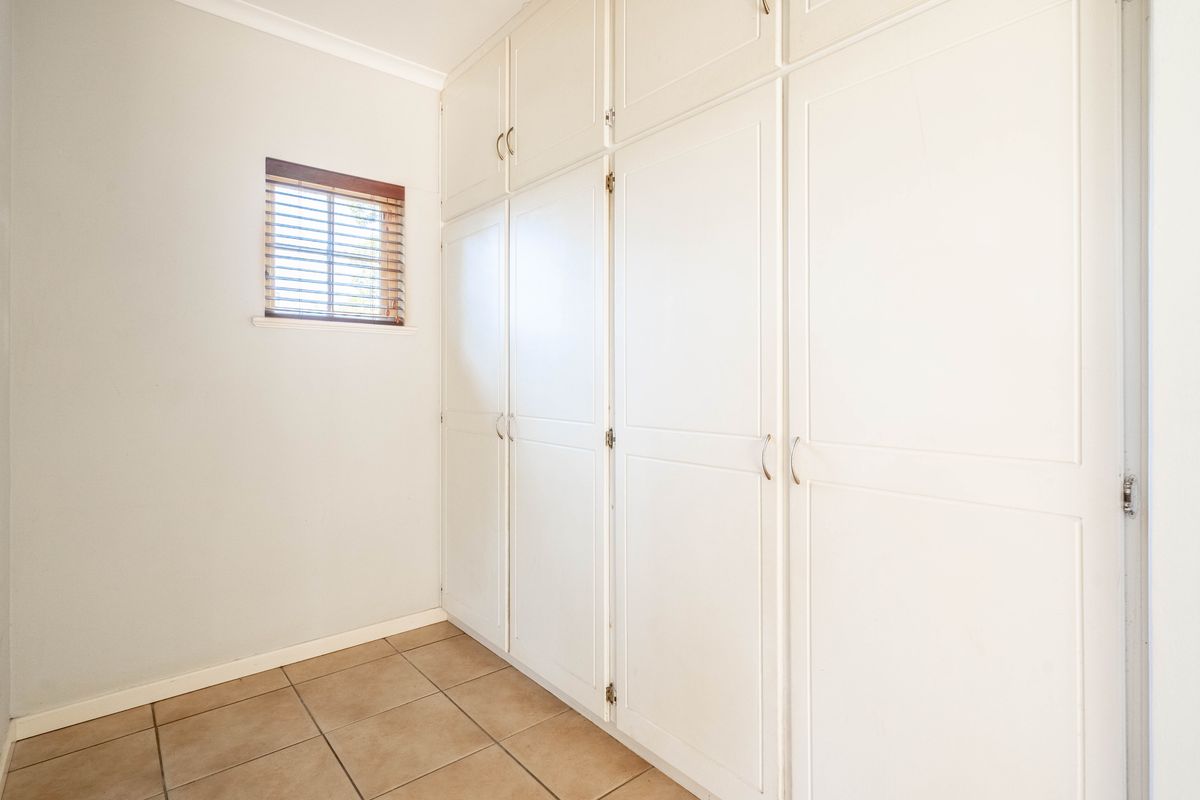 A clean, empty bedroom featuring extensive white built-in cupboards, neutral tiled flooring, and a small window with fitted blinds.