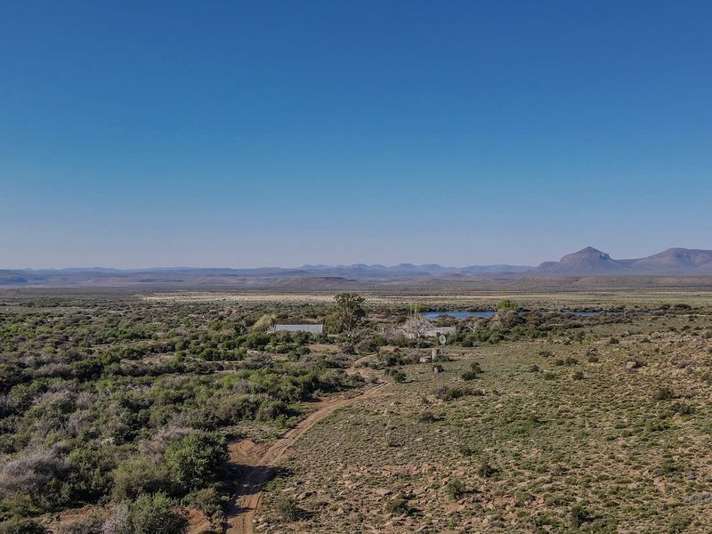 An aerial view of the expansive farm landscape featuring a windmill, a small dam, and surrounding natural vegetation under a clear blue sky.